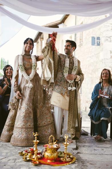 Photo of a young Indian couple in traditional Indian wedding attire-- actress Anjli Mohindra who played "The Rani" in recent Doctor Who spinoff the "Sarah Jane Adventures," and Sacha Dhawan, who played The Master against Jodi Whitaker's Doctor Who. On the ground in front of them are a variety of traditional Indian wedding dishes and objects. The couple both have one of their hands held up in the air and tied lightly together with a small piece of red rope.