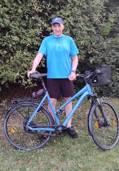 Ich stehe auf einer Wiese. Hinter mir ist ein Busch. Vor mir steht mein blaues Fahrrad. Es hat einen niedrigen Einstieg. Ich trage eine blaue Mütze und ein blaues Shirt. Außerdem eine kurze schwarze Hose und schwarze Schuhe.