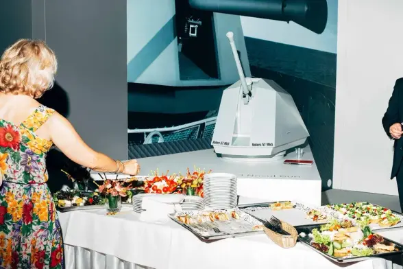 Das Foto zeigt ein kaltes Buffet, an  dem sich eine von hinten sichtbare Frau bedient. An einer Wand ist der Kanonenturm eines Kriegsschiffes zu sehen, der auch als Modell auf einem Tisch hinter dem kalten Buffet zu sehen ist.
