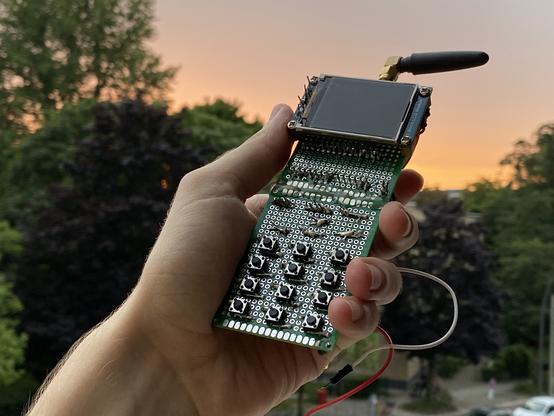 A "phone" consisting of a visible circuit board with buttons and a screen soldered onto. Wires are coming from the back some not soldered on to the phone. At the top is a antenna. The soldering job is exceptionally bad.