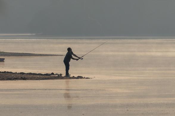 Fisherman in silhouette, standing on a bit of land extending into a lake, holding a fishing rod. Misty water in the foreground and background.