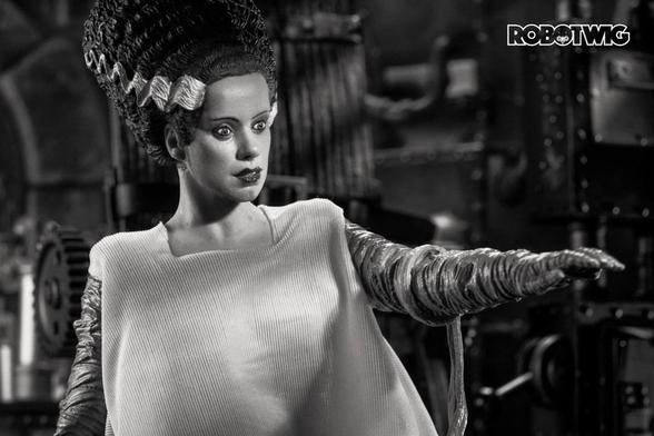 The Bride of Frankenstein extends her arm, looking at her bandages. Background is a lab setting