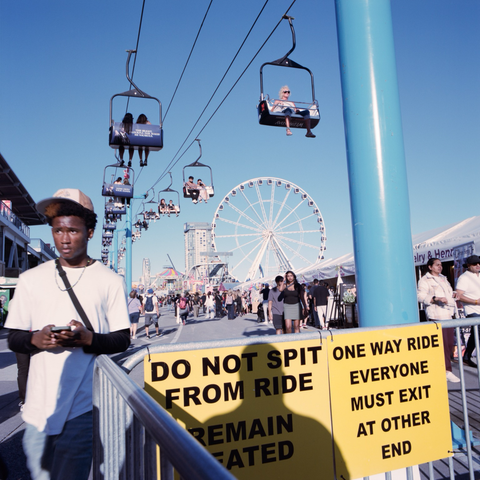 “Do not spit from ride. Remain seated.” warns a sign at one end of the SkyRide, an overhead chairlift that runs from one end of the main fairground to the other. People stroll around below with the midway in the distance.