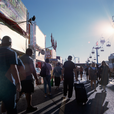 “Chunks o Chicken” and “Curly’s Fries”, people silhouetted by the afternoon sun that flares the end of the frame, SkyRide overhead.
