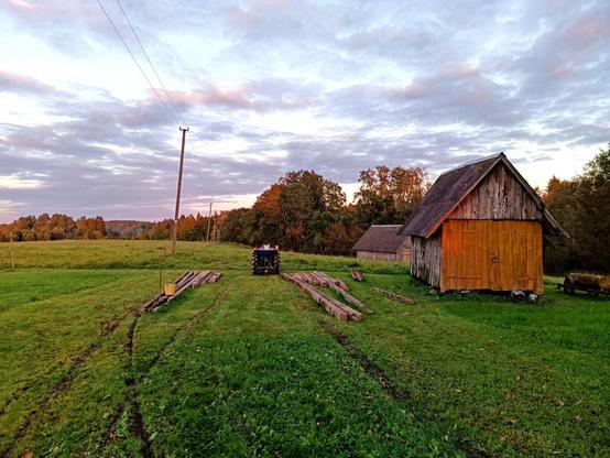 An area of mowed grass with a barn (and another barn behind it). Two lots of long concrete power pylons lay on the ground in various lengths and conditions. A small blue tractor is idling nearby after pulling them there. The grass has curving stripes of bare dirt from where the beams where dragged over it. The sun is low and the sky cloudy.
