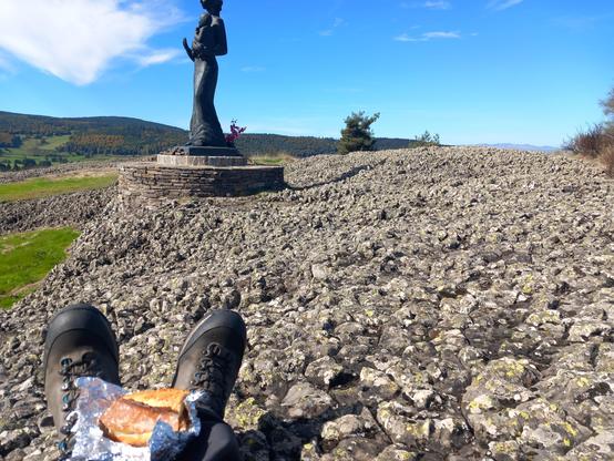 Au premier plan, un sandwich dans du papier d'aluminium posé sur mes pieds. Derrière, un terrain rocheux irrégulier avec quelques zones herbeuses sur les côtés. Dans le prolongement de mes pieds, une statue de vierge à l'enfant en bois. Au loin à droite, tout au fond, des crêtes rocheuses. Le ciel est clair avec quelques petits nuages blancs.