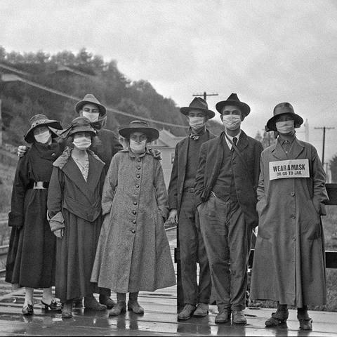 A black and white historical photograph featuring a group of seven individuals dressed in early 20th-century clothing, all wearing masks. One person holds a sign that reads, "WEAR A MASK OR GO TO JAIL." The setting is outdoors.