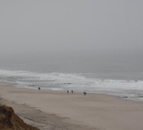 Blick aus erhöhter Position auf einen weitläufigen Strand, indem nur wenige, ganz kleine Menschenfiguren zu sehen sind. Deutlicher Wellengang. die Luft ist diesig