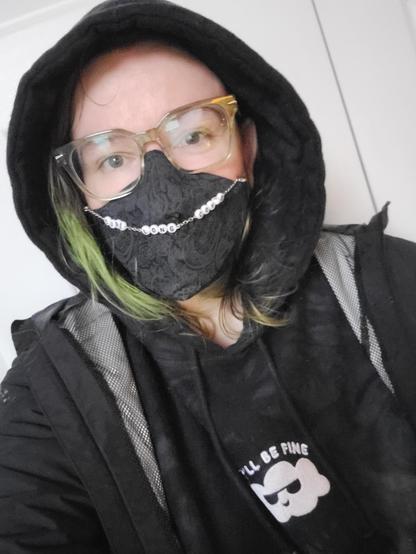 Photo of a white person with black and green hair, green glasses, black tie dye hoodie with the hood up, and black rain coat on showing off their 3M aura N95 mask cover in a black paisley with a mask chain that reads "cure long covid".