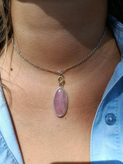 a silver pendant with a big oval pink stone and a smaller round green one. it has a silver chain. it's worn on a woman's bust