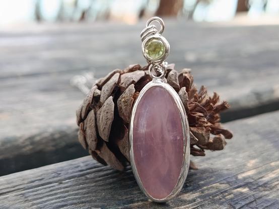 a silver pendant with a big oval pink stone and a smaller round green one. it has a silver chain. a pine cone for background