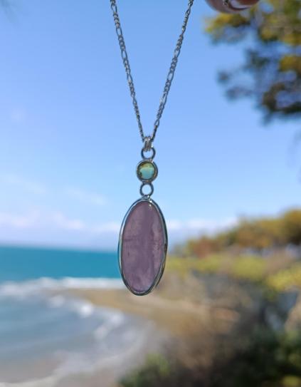 a silver pendant with a big oval pink stone and a smaller round green one. it has a silver chain. it's hanging in the air and in blurry background there is a beach