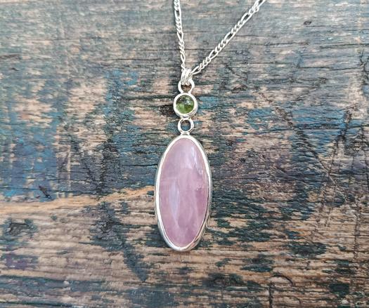 a silver pendant with a big oval pink stone and a smaller round green one. it has a silver chain. it's placed on piece of wood