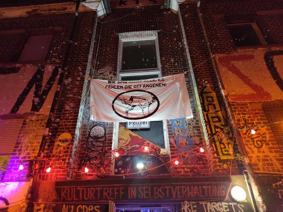 Blick von unten nach oben auf eine Hausfassade (Backsteinfassade) bei Nacht. Beleuchtet von kleinen Lampen. einige Fenster. Es handelt sich um die Fassade eines bunt bemalten Autonomen Zentrums. An der Fassade hängt ein weißes Banner mit der Aufschrift "Es fehlen die Gefangenen"