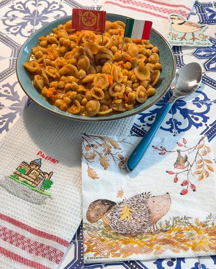 in Stillleben mit einem Teller Nudeln, einem Löffel und einer Serviette auf einem Tisch. Der Teller ist blau mit einem weißen Muster. Auf dem Teller befindet sich eine Portion orangefarbener Nudeln mit einer kleinen Gabel, die mit „Vegan“ beschriftet ist. Ein silberner Löffel liegt rechts vom Teller auf dem Tisch. Eine quadratische Serviette mit einem Muster aus orangefarbenen, gelben und braunen Bildern ist links vom Teller auf dem Tisch platziert. Auf der Serviette befindet sich eine Illustration eines Gebäudes mit der Aufschrift „PALERMO“ und der URL www.albinente.eu. Der Hintergrund ist ein Tisch mit einem blauen und weißen Muster.