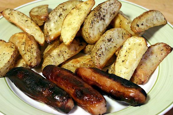 Three well done British style sausages on a plate, backed by a pile of oven roasted potato wedges with herby bits.