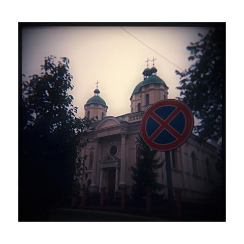 A square, low-fidelity analog photograph taken with a Holga camera, showing the facade and twin green domes of a church against a hazy, overcast sky. A large, circular red and blue 'No Stopping/Parking' traffic sign is prominently positioned in the right foreground, partially obscuring the building. Dark foliage frames the scene on the left and right, emphasizing the characteristic Holga vignette.
