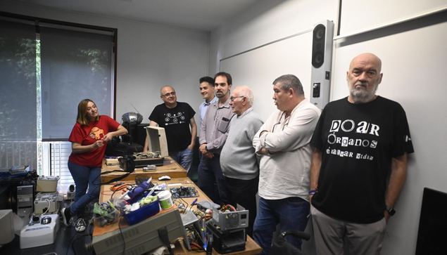 Voluntariado posando ante mesa longa chea de cachivaches en reparación, no Banco do Campus Industrial de Ferrol con membros das asociacións Alcer e CRS Narón. Foto de Emilio Cortizas de Diario de Ferrol