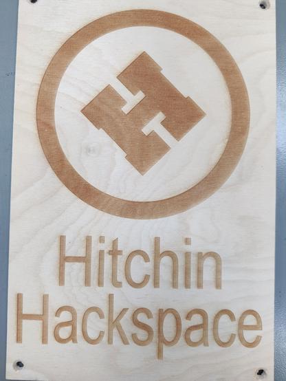 At the top of the image is the Hitchin Hackspace "H" logo in the centre of a circle. The H is at 45 degrees and the character has a square outline. The words "Hitchin Hackspace", along with the H in the circle, are laser engraved on to an A4 sized sheet of Birch plywood.
It should be at the pub, to help identify us, providing one of our members remembers to bring it!🤦‍♂️