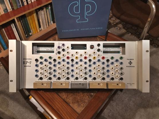 An electronic device in standard 19"-rack size on a small table. The device has many connectors on the front in a hexagonal grid. Lettering on the front says "Operational RP-F Manifold" and "Philbrick Researches".