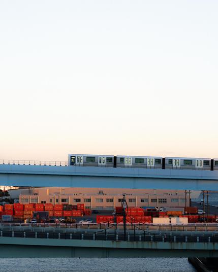 Train traveling on an elevated railway bridge during sunset, with industrial buildings and stacked orange shipping containers below. The scene includes a large expanse of the sky and water in the foreground, creating a layered composition.
