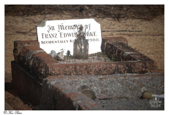 A close up photograph of a historically significant, simple grave marker at the Kingston on Murray Cemetery.  A small white stone plaque, leaning slightly, is clearly readable and marks the grave of Franz Edwin Obke, noting he was "ACCIDENTALLY KILLED 1908."  The grave is outlined by a rough, reddish-brown brick border, and in the center rests an old, cloudy glass jar holding a withered flower, signifying a recent or ongoing tribute.  The surrounding ground is sparse and dirt-covered, emphasizing the isolated, historic nature of the resting place. Signed Kev Peirce.