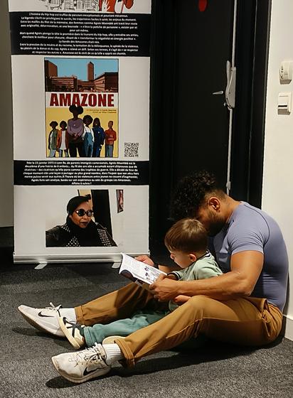 Photo. numérique en format portrait, avec au premier plan un homme très musclé des bras assis par terre sur la moquette grise. Entre ses jambes écartées, son fils est assis à lire une BD tenue par son père.
A l'arrière-plan, un kakemono (affiche haute tenue par un support) présente le parcours d'Agnès Maemblé, avec du texte blanc sur fond noir, une photo avec des lunettes de soleil, le dessin de son groupe de rap avec 6 femmes et une photo d'un quartier moderne (la Part-Dieu à Lyon).