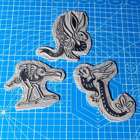 three grey carved linoblocks laid out on a blue cutting mat. they are strange little critters inspired by medieval marginalia: a long legged bird like creature with a curling tail and very long beak. a creature with a bunny's head, upraised birds wings and forked lizards tail, and a flying snake like creature with feathery wings, a large leaf like structure at the end of their tail and a round furry head and sharp teeth.