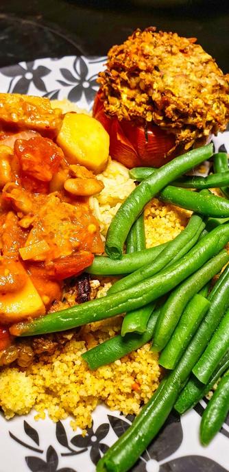 A plate with roasted stuffed tomato; Walnut and Oat burger, topped with spiced couscous, rice, and Pinto bean & Seitan Chilli, served with Green beans.