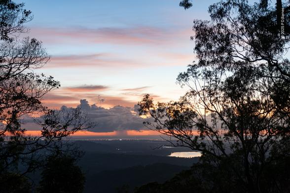 A gentle pink sunrise with soft, high clouds is contrasted by a low heavy cumulus not far above the sea, with a very obvious downpour falling from one section into the ocean. The sky overhead is blue and clear, and the  plains below are still dark save for the shining patch of a large dam. A line of coastal lights marks the boundary between land and sea, and the whole image is heavily framed left and right by tree silhouettes. The rain will pass, and so will the sunrise, life is change, embrace the journey.