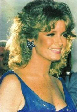 A candid 1986 portrait of Deidre Hall, best known for her role as Marlena Evans on Days of Our Lives, taken at the 33rd Annual SHARE Boomtown Party in Los Angeles. She wears a vibrant blue sleeveless dress with a deep neckline and a star-shaped earring adorned with small white dots. Her shoulder-length light brown hair is softly layered and styled in voluminous curls typical of the 1980s. She is smiling warmly and looking slightly away from the camera. The background is dark, suggesting an indoor event setting. The image captures a moment of elegance, warmth, and nostalgic glamour, reflecting Deidre’s iconic status in daytime television.