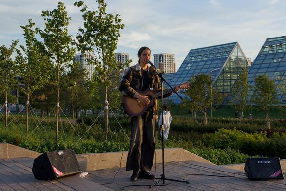 The image depicts a man with long straight black hair standing on a stairs in a park, playing a guitar and singing into a microphone. He is dressed in a black jacket and pants, with his hands positioned on his guitar as he performs. The stage is set against the backdrop of several building, which appears to be made of glass and steel. This modern structure contrasts with the natural setting of trees visible in the background.