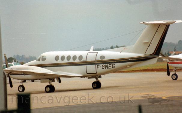 Side view of a twin-engined BizProp parked facing to the left.
The plane is mostly white, with a black and gold stripe running along the body, sweeping up into the rear of the tail.
The black registration "F-GNEG" is on the lower rear fuselage, just aft of the main door.
Other, smaller, planes can be seen in the background, on either side.
Trees stretch across the frame in the distance, under flat grey sky.