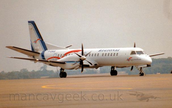 Side view of a twin propellor-engined airliner parked facing to the right.
The plane is mostly white, with diagonal dark blue stripes with light blue piping along the leading and trailing edges of the tail, running down to loop under the rear fuselage. Overlaid with a stylised red flying bird over a blue ord.
A larger version of the red bird logo adorns the rear fuselage, as well as a smaller version with small "Regional" titles under the cockpit.
There are larger "Regional" titles on the upper forward fuselage, and the black registration "F-GMVB" on the upper rear.
Thin dark and light blue stripes run along the length of the engine pods in front of the wings.
Sandy-grey concrete apron marked with yellow lines for planes to follow fills the foreground, with trees in the distance, under grim, hazy sky.