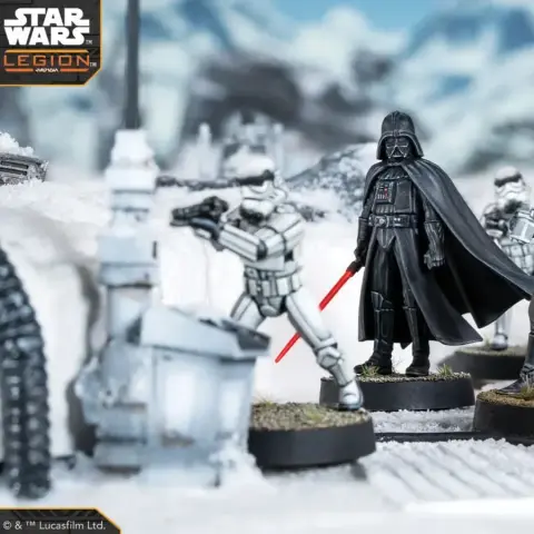A detailed scene features Darth Vader standing prominently with his red lightsaber drawn, dressed in his iconic black armor and cape. Flanking him are two Stormtroopers, one aiming a blaster, set against a snowy landscape with scattered debris and structures. The miniatures are mounted on circular bases, showcasing their intricate designs and the dynamic pose of the characters.