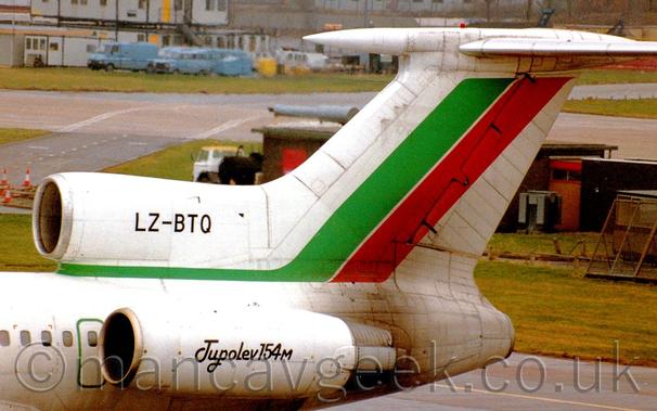 The rear fuselage of a 3 engined jet airliner, with the engines mounted on the top and sides of the rear fuselage, parked facing to the left.
The plane is mostly white, with green stripe running along the top of the rear fuselage under the centre engine, sweeping diagonally up into the white tail, where it is joined by a diagonal red stripe.
The black registration "LZ-BTQ" is on the centre engine intake, while the side mounted engine has black text "Tupolev 154M".
A dark brown brisk building in the background is partially hidden by the tail, with a taxiway beyond that, and blue airport vans parked in front of some grey pre-fabricated buildings in the distance.