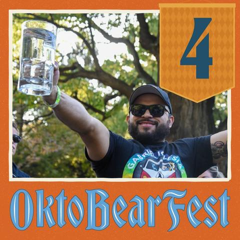 The image shows a man wearing a baseball cap and sunglasses, standing in front of a large orange banner with the word "Oktoberbearfest" written on it. The man is holding a glass of water, which he is holding up in the air to the camera. In the background, there are additional people present, but their features are not as clearly visible.