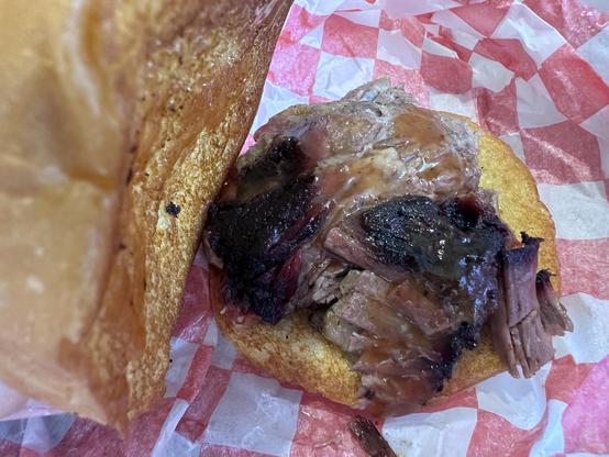 A close-up of a sandwich featuring tender pulled barbecue meat on a toasted bun, resting on a red and white checkered paper. The meat is dark and saucy, indicating a rich flavor.