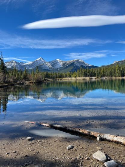 Serene mountain lake scene with evergreen trees, snow-capped peaks, blue sky, and calm water reflecting the landscape.