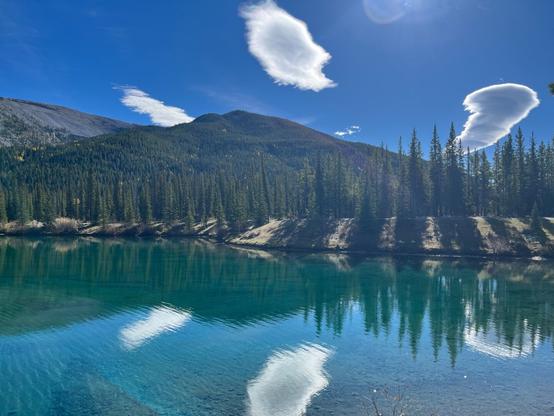 Clear turquoise lake with reflections of trees and mountains under a blue sky with scattered clouds.