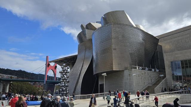 The exterior of the Guggenheim Museum, all silvery shiny plates covering the walls that are winding and bending in various sleek curves around many big elements, not a singular 90 degree angle in sight and barely any straight lines. People are walking on sandy coloured paths around the building. An art installation made of many huge silver balls is to the left of the museum. In the background is a bridge with a comic like red gigantic bridge pillar. Between grey clouds the blue sky is visible.