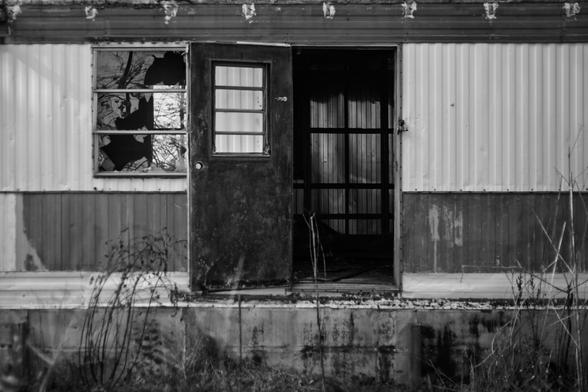 Black and white photo of an abandoned trailer with a rusted metal door hanging open. The window beside it is shattered, and tall dry weeds grow in front. The inside is dark and empty, revealing wooden slats and shadows beyond the doorway. The structure’s corrugated metal siding is weathered and peeling.