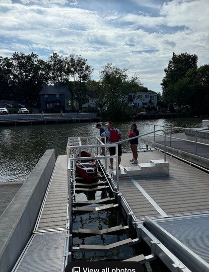 image of a paddling boat in an adaptive dock. The boat (kayak-like, but more open body) is just above the water in struts which looks like it holds it steady. and there are grab bars along the boat size and multiple metal contraptions I don't understand overhead but look weight bearing, and perhaps could be used for attaching the wheelchair transfer I saw on their website? There's also a set of metal steps with a sturdy railing, perhaps as an alternative to the ramp? Three humans are about to get in the boat. The canal is somewhat backlit, wide and textured. the sky is partly cloudy with whispy clouds.
