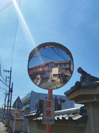 Just outside the temple is a pole with a round traffic mirror. Behind it is the temple grounds wall and a blue sky. In the mirror, if one looked closely, one would see one of the two Buddha statues that were erected on the opposite side of the road. The sun just outside the shot in the upper left hand corner gives off a bit of lens flare.