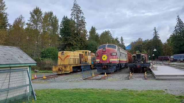 several trains on storage tracks. A torn down engine. An old EMD F7 red and yellow locomotive number 1404. yellow and orange yard switcher locomotive. trees and clouds in the background