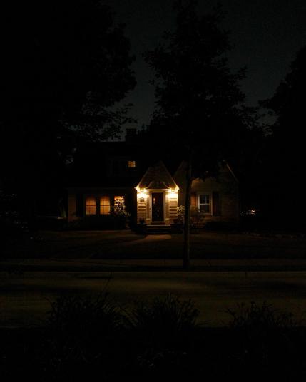 Color photograph looking across a residential street at night at the front of a house with bright porch lights bracketing a dark brown door, the rest of the face of the house darkened into silhouette around the door, roofline blending with darkened silhouettes of trees around the house under a dimly glowing night sky