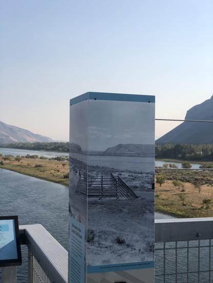 Photo of an info sign with a historic photo that nearly lines up with the river and hillside behind it.
