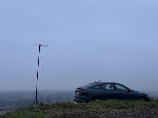 Das Bild zeigt einen dunklen, möglicherweise blauen, Audi e-tron auf einer kleinen Anhöhe. Neben dem Auto steht ein Stativ mit einer Antenne. Im Hintergrund ist ein nebliger Ausblick auf eine Landschaft zu sehen. Der Himmel ist bedeckt und grau.