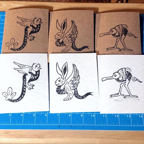 Six block print cards, three parchment patterned and three kraft, with three different prints on them, they are strange little critters inspired by medieval marginalia: a long legged bird like creature with a curling tail and very long beak. a creature with a bunny's head, upraised birds wings and forked lizards tail, and a flying snake like creature with feathery wings, a large leaf like structure at the end of their tail and a round furry head and sharp teeth.