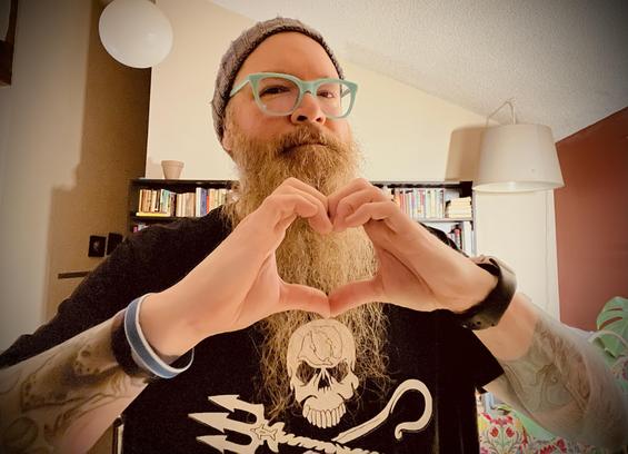 Photo of me in front a bookshelf inside a house. I’m holding up my hands in the shape of a heart and looking into the camera with an expressionless face. I’m wearing a knit hat, sea foam green glasses, and a black Sea Shepherd t-shirt. I have a long beard and have two full tattoo sleeves.